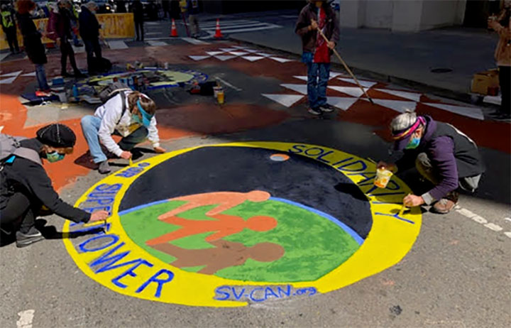 Three people working on a street painting that shows three different colored stick figures holding hands in front of a black, green, and blue. background. The figures are surrounded by the words Solidarity is our superpower, SV-CAN.org" in blue writing on a yellow background.